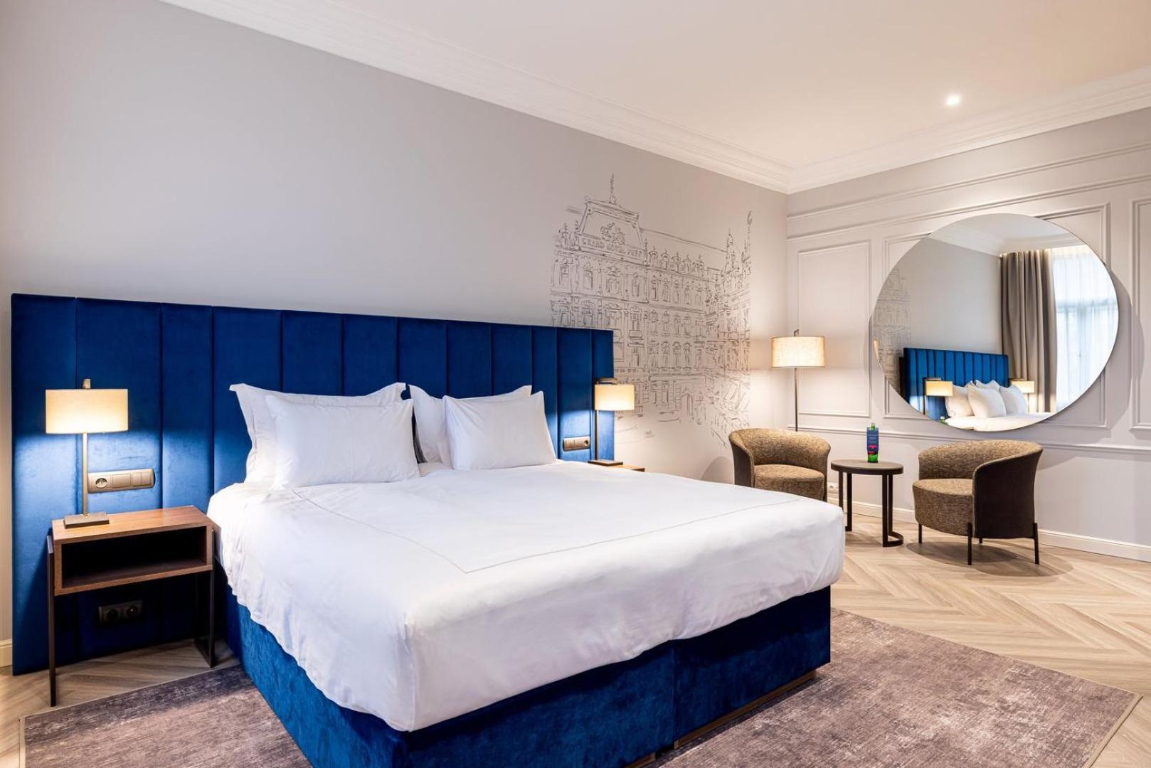 Grandhotel Pupp Karlsbad Buitenkant foto The photo shows a modern hotel room featuring a large double bed with white linens and a blue upholstered headboard. The walls are light gray, and there is a decorative mural in the background. Two bedside lamps are on either side of the bed. In the 