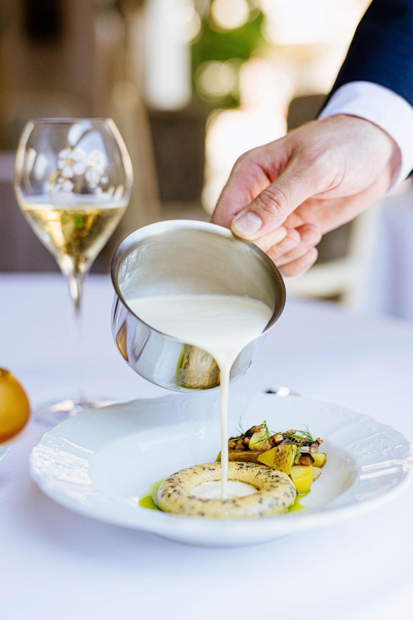 Grandhotel Pupp Karlsbad Buitenkant foto The photo shows a hand pouring a creamy sauce from a small stainless steel container over a plated dish, which appears to include vegetables and possibly pasta. In the background, there's a glass of sparkling wine, suggesting a fine dining setting. T