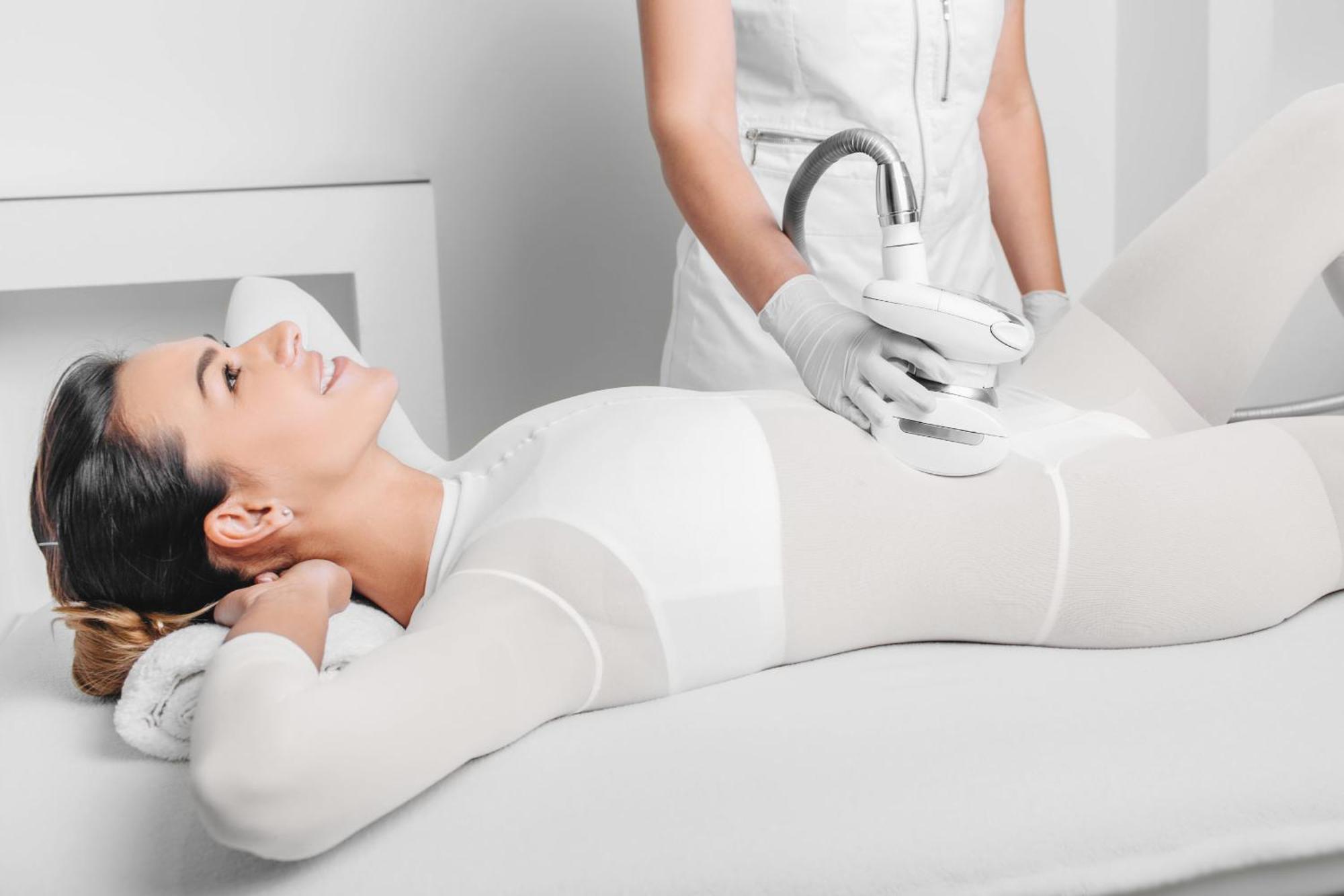 Grandhotel Pupp Karlsbad Buitenkant foto The photo depicts a woman lying on a treatment table, appearing relaxed and smiling. She is wearing a form-fitting white outfit, likely designed for a cosmetic or wellness procedure. A technician, dressed in a white outfit and gloves, is applying a d