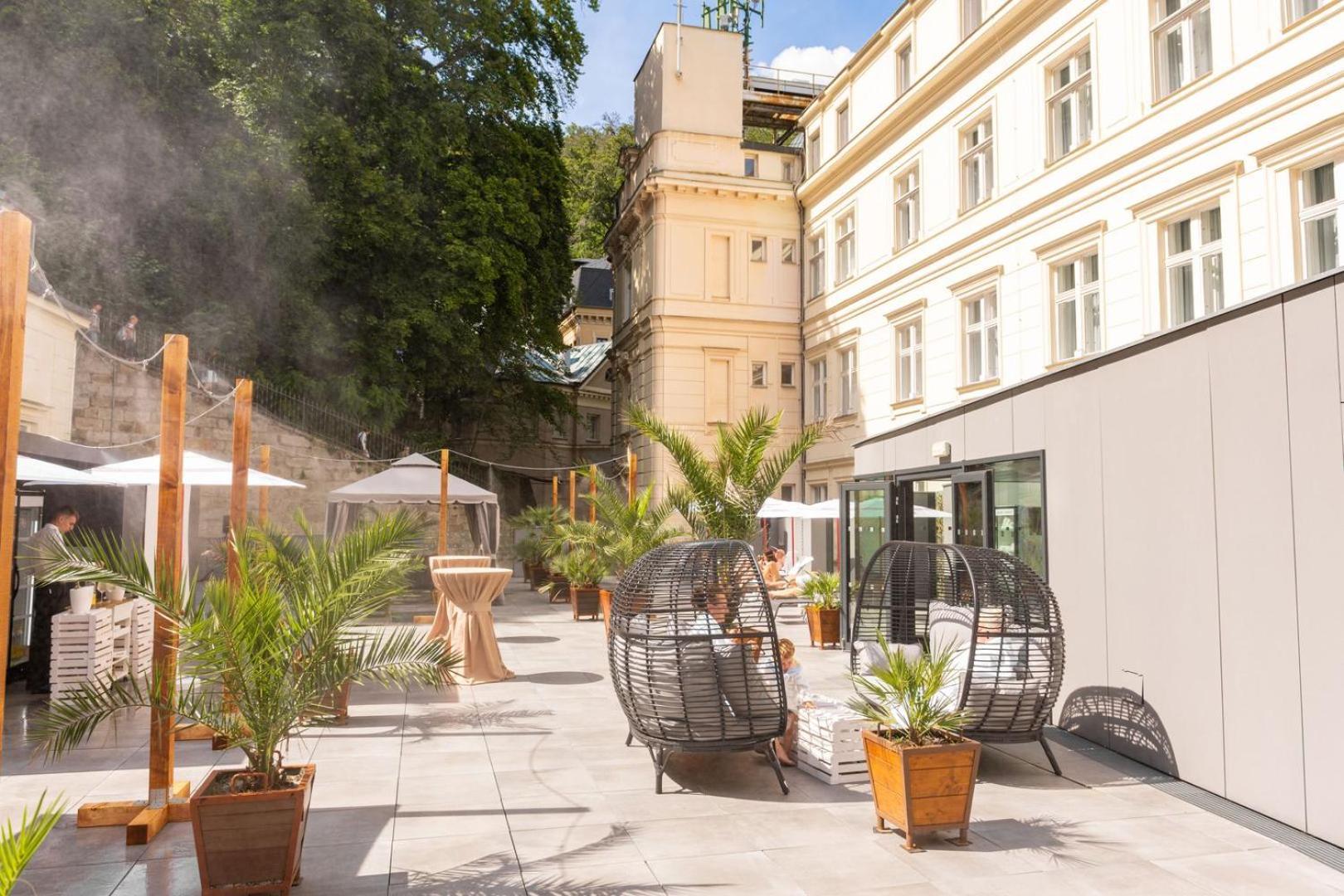 Grandhotel Pupp Karlsbad Buitenkant foto The photo shows a stylish outdoor area with a mix of modern and classic architecture. There are several large potted plants, likely palm trees, which add a tropical feel to the space. Two round, open wicker chairs are placed on a paved surface, provi