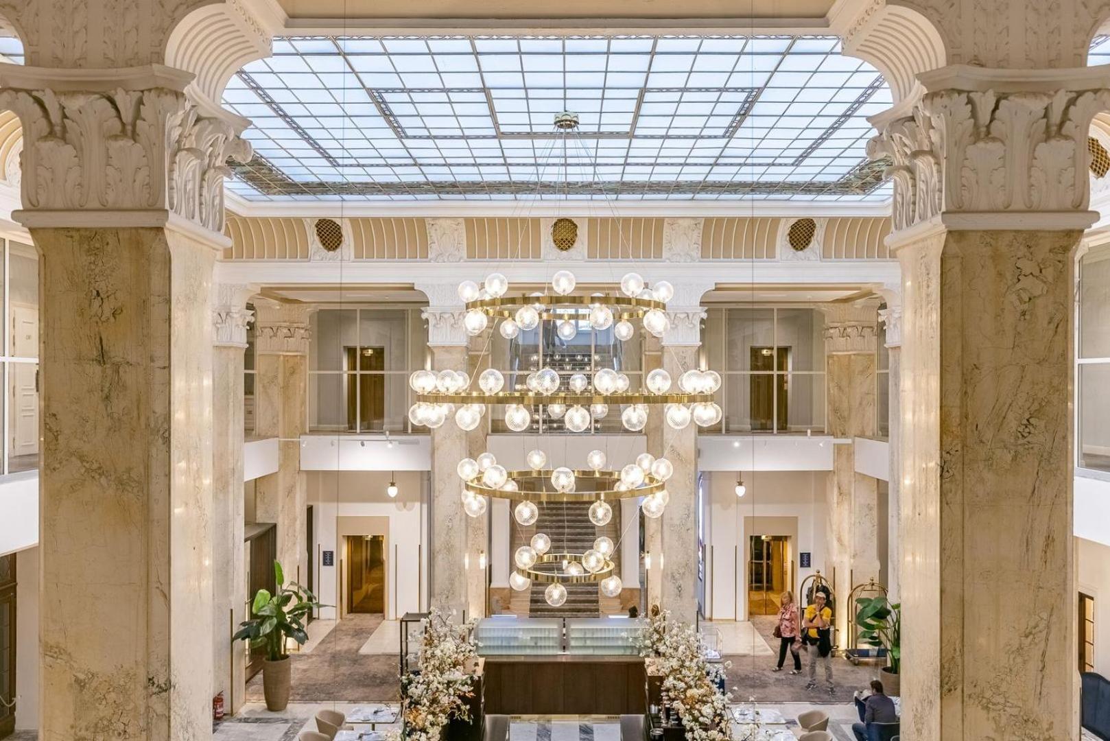 Grandhotel Pupp Karlsbad Buitenkant foto The photo depicts an elegant hotel lobby characterized by its spacious and luxurious design. Large marble columns rise to support a ceiling with a glass skylight, allowing natural light to illuminate the area. A central chandelier made of numerous ro